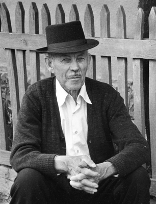 Idős férfi kalapot visel, és egy kerítés előtt ül, kezei összekulcsolva.