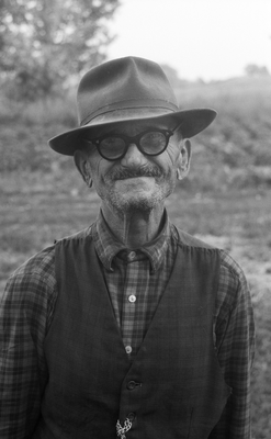 Idős férfi kalapban, kockás ingben és mellényben, háttérben természetes környezet