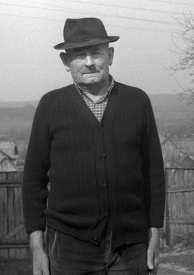 Idős férfi kalapot visel, pulóverben áll egy vidéki környezetben, háttérben kerítés és falu látható.