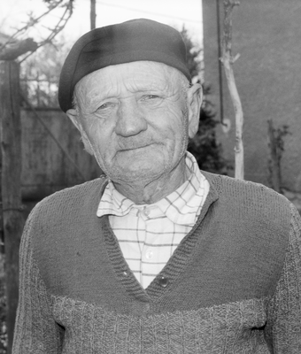 Idős férfi kockás ingben és kötött pulóverben, kalapot visel, a háttérben kert látható.