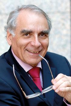 Idős férfi öltönyben, piros nyakkendővel és csíkos inggel, kezében szemüveget tart.