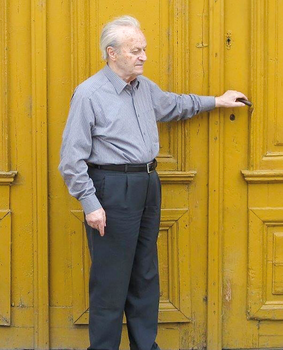 Idős férfi áll egy sárga faajtó előtt, kezével a kilincset fogva.