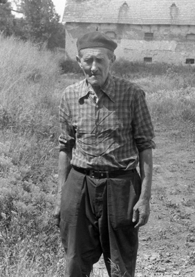 Idős férfi áll egy régi épület előtt vidéki környezetben, mezőgazdasági területen.