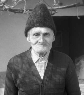 Idős ember hagyományos ruhában és sapkában, háttérben faágak és épület.