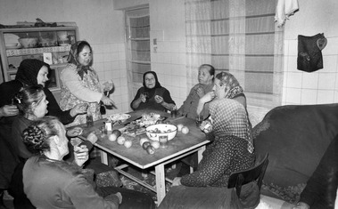 Idős emberek ülnek egy asztal körül, gyümölcsök és italok az asztalon, otthonos szoba háttérben.