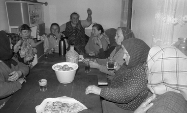 Idős emberek ülnek egy asztal körül, italokat fogyasztanak, gyümölcsök és sütemények az asztalon.