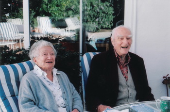 Két idős ember ül egy teraszon, háttérben kerti bútorok és növények láthatók.