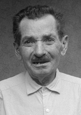 Idős férfi világos ingben, mintás háttérrel, homályosított arccal