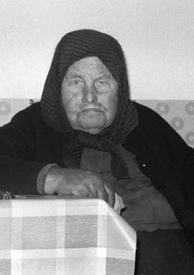 Idős nő fejkendővel ül az asztalnál, fekete-fehér fénykép.