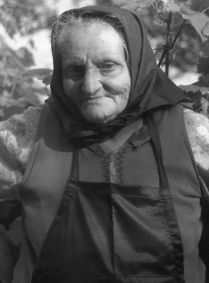 Idős nő fejkendőt visel és kötényt hord, háttérben szőlőlevelek láthatók.