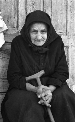 Idős nő fekete ruhában ül egy régi faajtó előtt, kezében egy botot tartva.