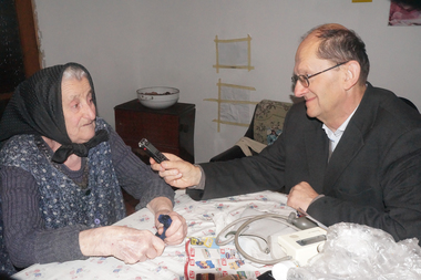 Idős nő és férfi ülnek egy asztalnál, a férfi diktafont tart a nő felé, az asztalon vérnyomásmérő és újságok.