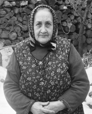 Idős nő hagyományos mintás ruhában áll, háttérben farakás látható.