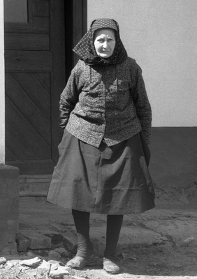 Idős nő hagyományos ruhában áll a ház bejárata előtt, kendőt viselve.