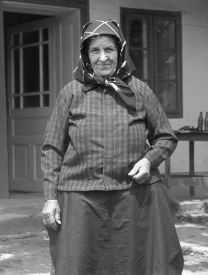 Idős nő hagyományos ruhában áll egy ház előtt, kendőt viselve.