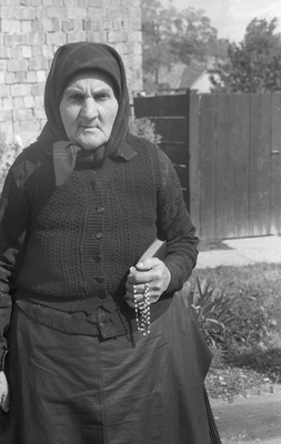 Idős nő hagyományos ruhát visel, kezében rózsafüzér, háttérben kőfal és kert.