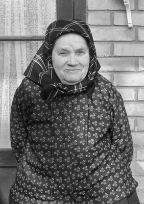 Idős nő hagyományos ruhában, kendővel a fején, téglafal előtt állva.