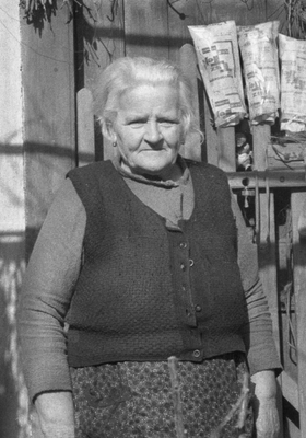 Idős nő hagyományos ruházatban áll egy fa kerítés előtt, háttérben papírcsomagok lógnak.