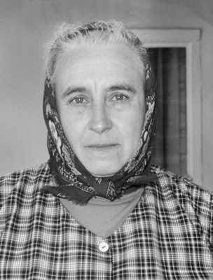 Idős nő kockás ruhát visel, fején kendő, fekete-fehér fotó.