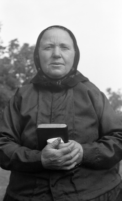 Idős nő fekete ruhában, kezében könyvvel, kültéri környezetben