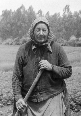 Idős nő fejkendőben, mezőgazdasági szerszámot tartva egy mezőn.