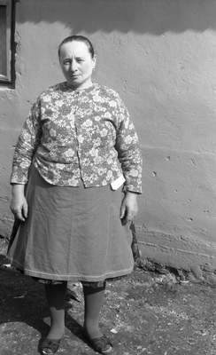 Idős nő virágmintás kardigánt és szoknyát visel, háttérben egy egyszerű fal.