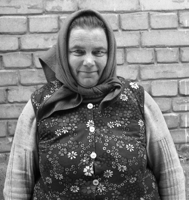 Idős nő virágmintás mellényben és kendővel, háttérben téglafal.