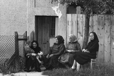 Öt idős nő hagyományos ruhában ül egy ház előtt, kerítés mellett, beszélgetnek és pihennek.