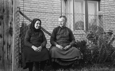 Két idős nő ül egy kerítés előtt egy vidéki ház udvarán, háttérben téglafal és ablak látható.