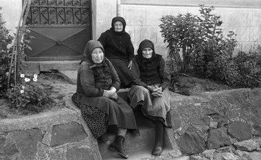 Három idős nő ül egy lépcsőn, fejkendőt viselnek, hagyományos ruházatban, háttérben kőfal és növények.