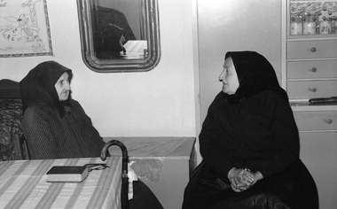 Két idős nő ül egy szobában, egyikük kezében rózsafüzér, az asztalon könyv és szemüveg.