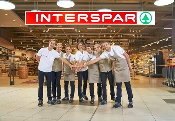 Interspar áruház dolgozói csapatmunka Interspar áruház bejárata, ahol dolgozók csoportosan állnak, kezüket összetéve, csapatmunkát szimbolizálva.