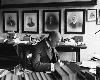 Férfi dolgozik egy íróasztalnál, körülötte könyvek és papírok, portrék a falon.