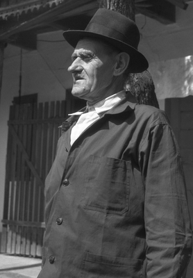 Kalapot viselő férfi áll egy fa előtt, háttérben kerítés és épület.