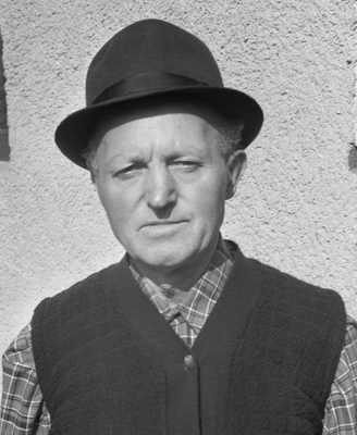 Kalapot viselő személy kockás ingben és kötött mellényben, háttérben fal.