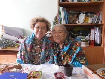 Két ember ül egy asztalnál, színes kendőbe burkolózva, étellel és itallal körülvéve, háttérben könyvespolc.