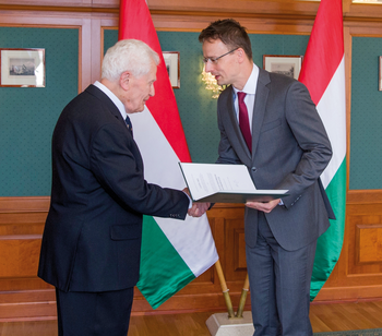 Két férfi kezet fog, egyikük oklevelet tart, háttérben magyar zászlók.