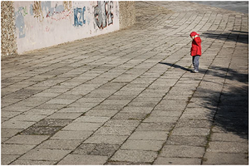 Kisgyermek térköves területen graffitivel díszített fal előtt Kisgyermek piros kabátban áll egy nagy térköves területen, háttérben graffitivel díszített fal.