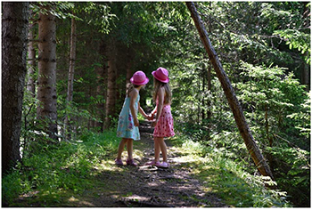 Két kislány rózsaszín kalapban az erdőben Két kislány rózsaszín kalapban és nyári ruhában kézen fogva áll egy erdei ösvényen.