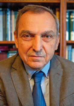 Könyvespolc előtt álló öltönyös férfi, akinek az arca el van homályosítva.