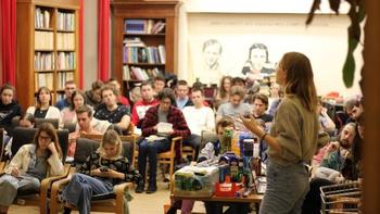 Könyvtári előadás termékbemutatóval Egy nő előadást tart egy könyvtárban, különböző termékekkel az asztalon, közönség előtt.