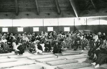 Közösségi gyűlés takarókkal egy fedett helyen Csoport ember takaróba burkolózva ül egy fedett helyen, valószínűleg egy közösségi eseményen vagy gyűlésen.