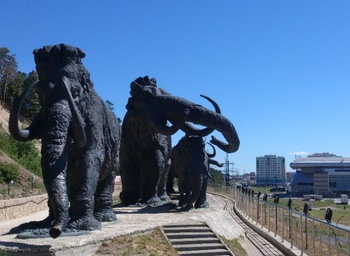 Mamut szobrok egy szabadtéri kiállításon Mamut szobrok láthatók egy szabadtéri kiállításon, városi háttérrel.