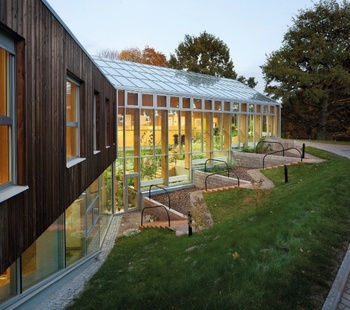 Modern üvegezett épület domboldalon, növényekkel és fa burkolattal.