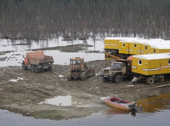 Munkagépek és lakókocsik sáros területen, csónak a vízparton, háttérben erdő.