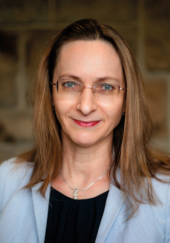 Nő világoskék blézerben, fekete felsővel és ezüst nyaklánccal, barnás fal előtt