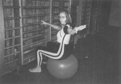 Egy nő tornateremben edz, gimnasztikai labdán ülve, karjait oldalra kinyújtva.