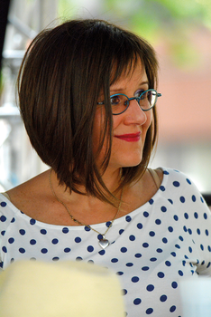 Nő pöttyös ruhában és nyaklánccal Nő pöttyös ruhában, nyaklánccal, rövid hajjal, háttérben zöld növények.