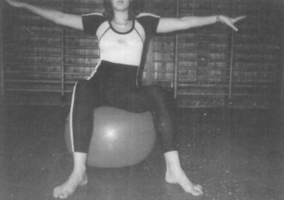 Egy nő ül egy fitnesz labdán egy tornateremben, karjait oldalra kinyújtva.