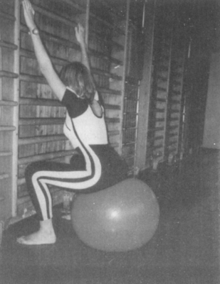 Egy nő gimnasztikai labdán ülve nyújtózkodik a bordásfal felé egy tornateremben.